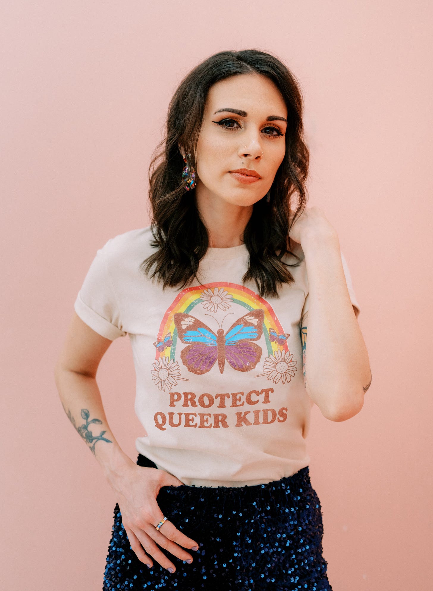 A photo of Shohreh from the waste up wearing navy blue sequined pants and a tucked in tshirt with a rainbow over a butterfly that reads, "Protect Queer Kids"