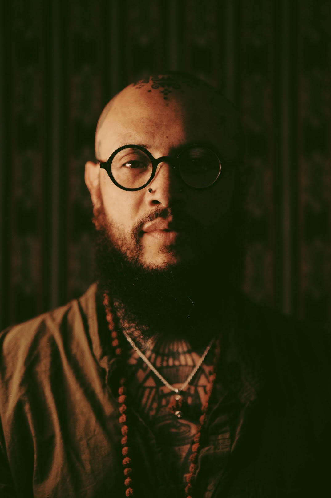 bald brown skin man with a geometric tribal tattoo on his head, with 2 nose piercings on either side. He is wearing black circular glasses