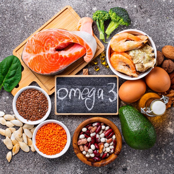 salmon, flax seeds, spinach, pumpkin pepitas, lentils, beans, avocado, eggs, walnuts, shellfish and broccoli arranged on a counter around a chalkboard that reads Omega 3