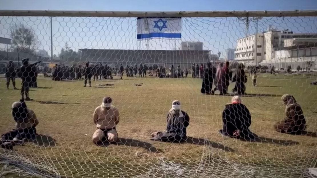 A still from a video appears to show Palestinian women and men blindfolded with their hands tied behind their backs as they sit on the grass in front of a soccer goal in the stadium.