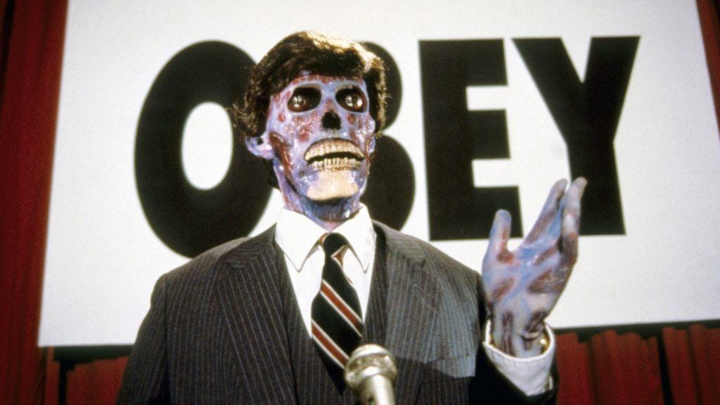 Screencap from They Live of an unmasked alien impostor speaking at a political rally in front of an "OBEY" sign.
