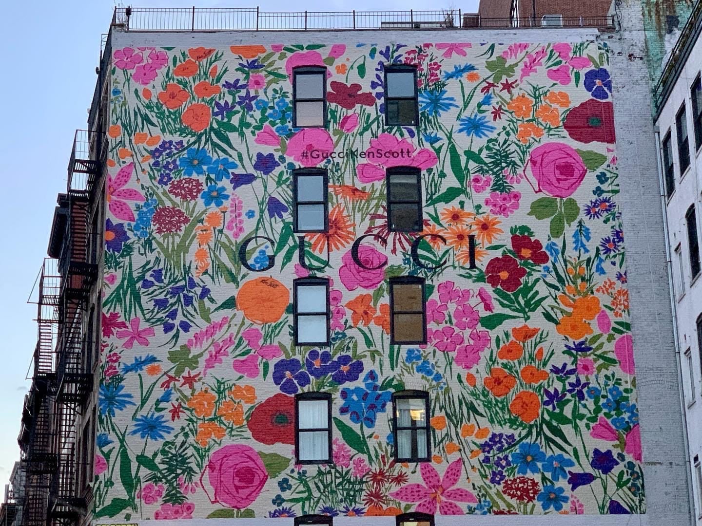 The side of a multi-story brick building with windows and a mural of all kinds of flowers with the word GUCCI centered.