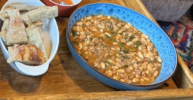 A wooden tray, with two dishes on it. One is a large blue flat dish, filled with stew. It's an orange brown colour, with flecks of green, white beans, and chunks of fish. Next to it is a smaller white dish filled with crusty bread.