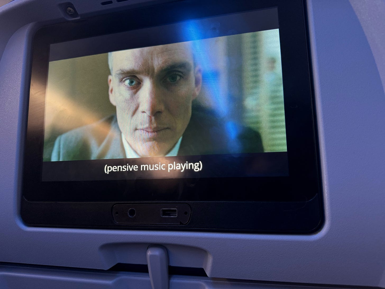 Photograph of an aircraft seatback with a video screen playing the movie 'Oppenheimer' with the words (pensive music playing).