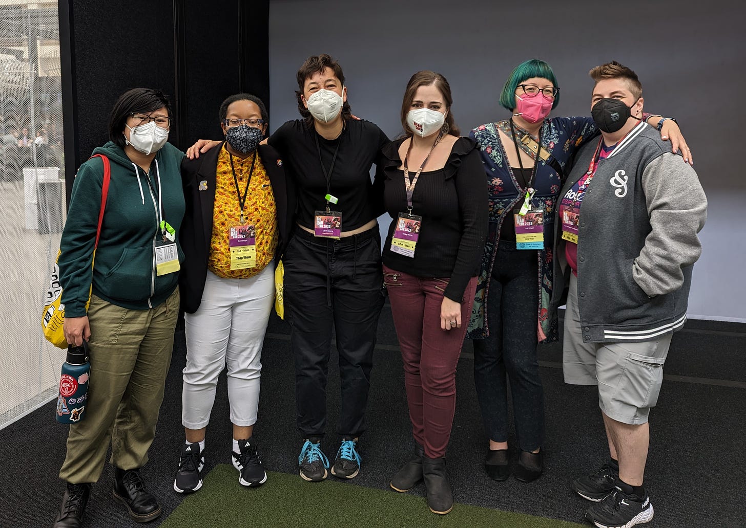 A group of six creatives wearing masks stand side by side in front of a gray wall.