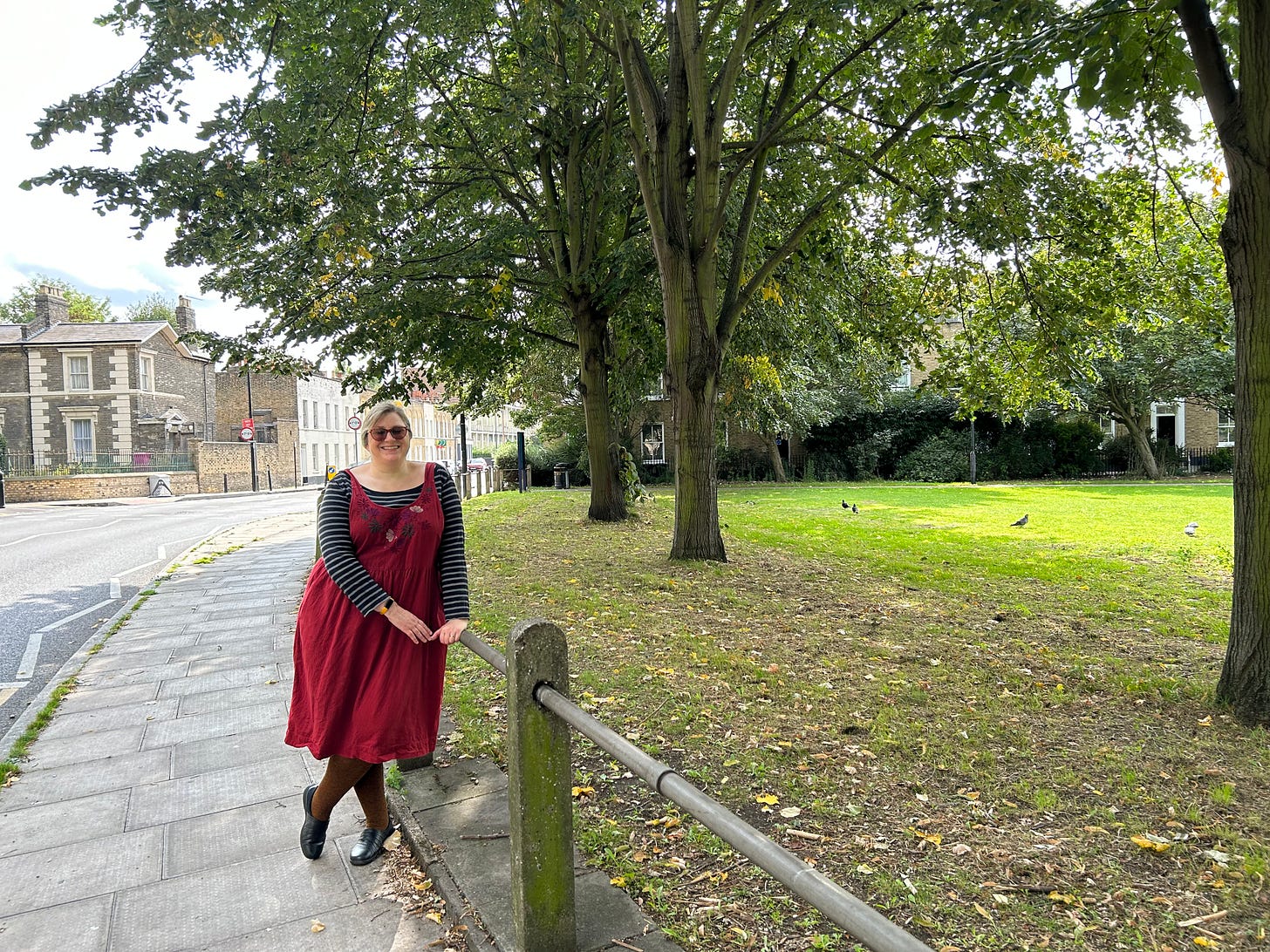 A white woman in a red dress stands leaning on a rail next to a grassy area with large trees, by the roadside