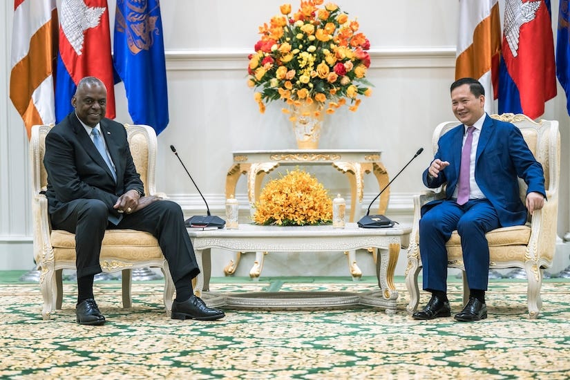 Two smiling people in business attire sit in chairs opposite one another with flags in the background.