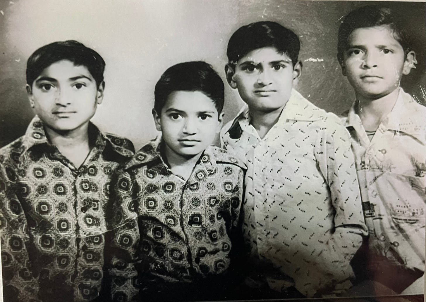 A black-and-white photo of a group of four Indian boys, each wearing a patterned long-sleeved shirt. 