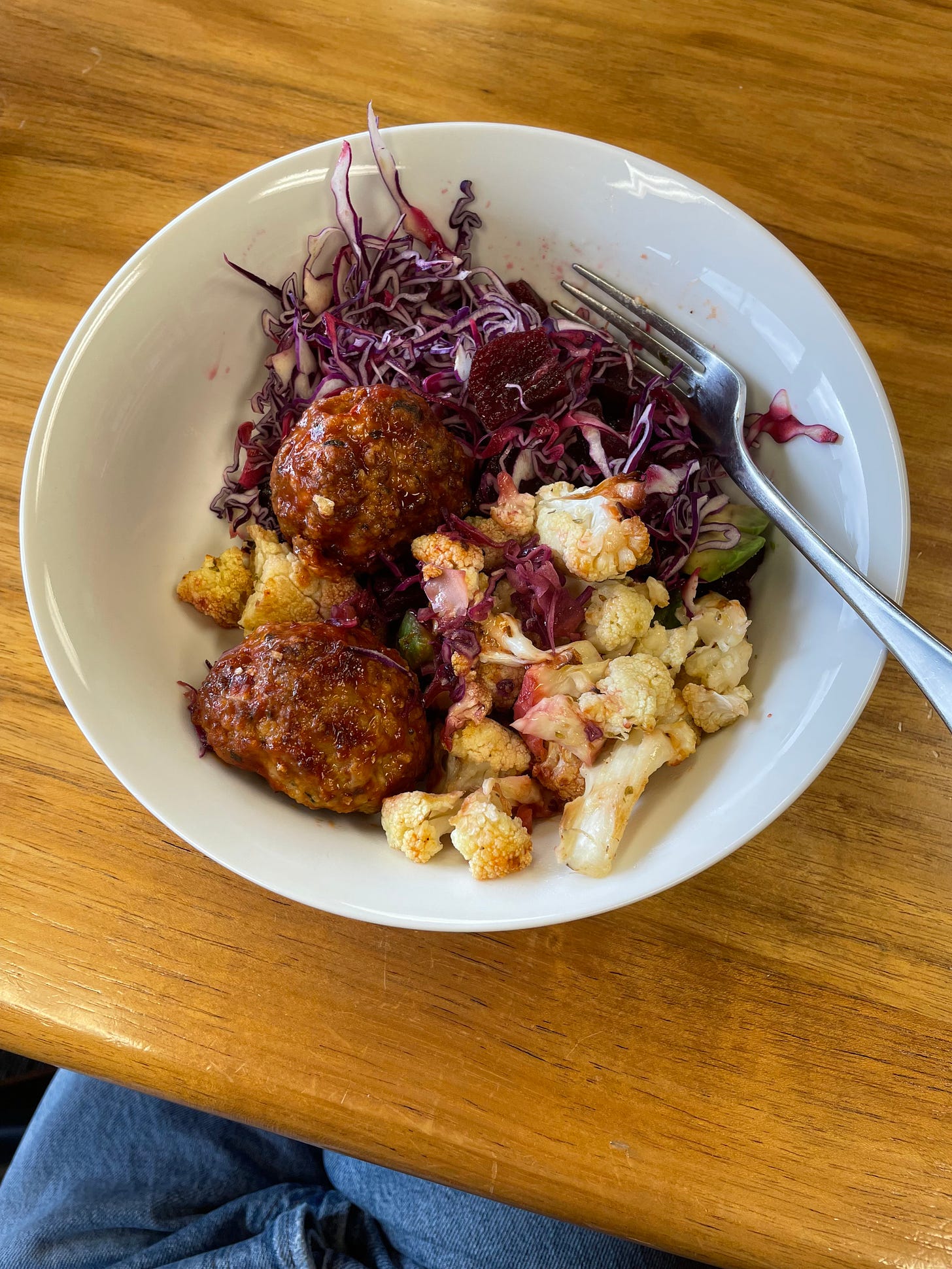 White bowl filled with cheesy cauliflower bites, shaved cabbage, beetroot and avocado, and two sticky chicken meatballs. Photographed on a wooden table in an office kitchen.