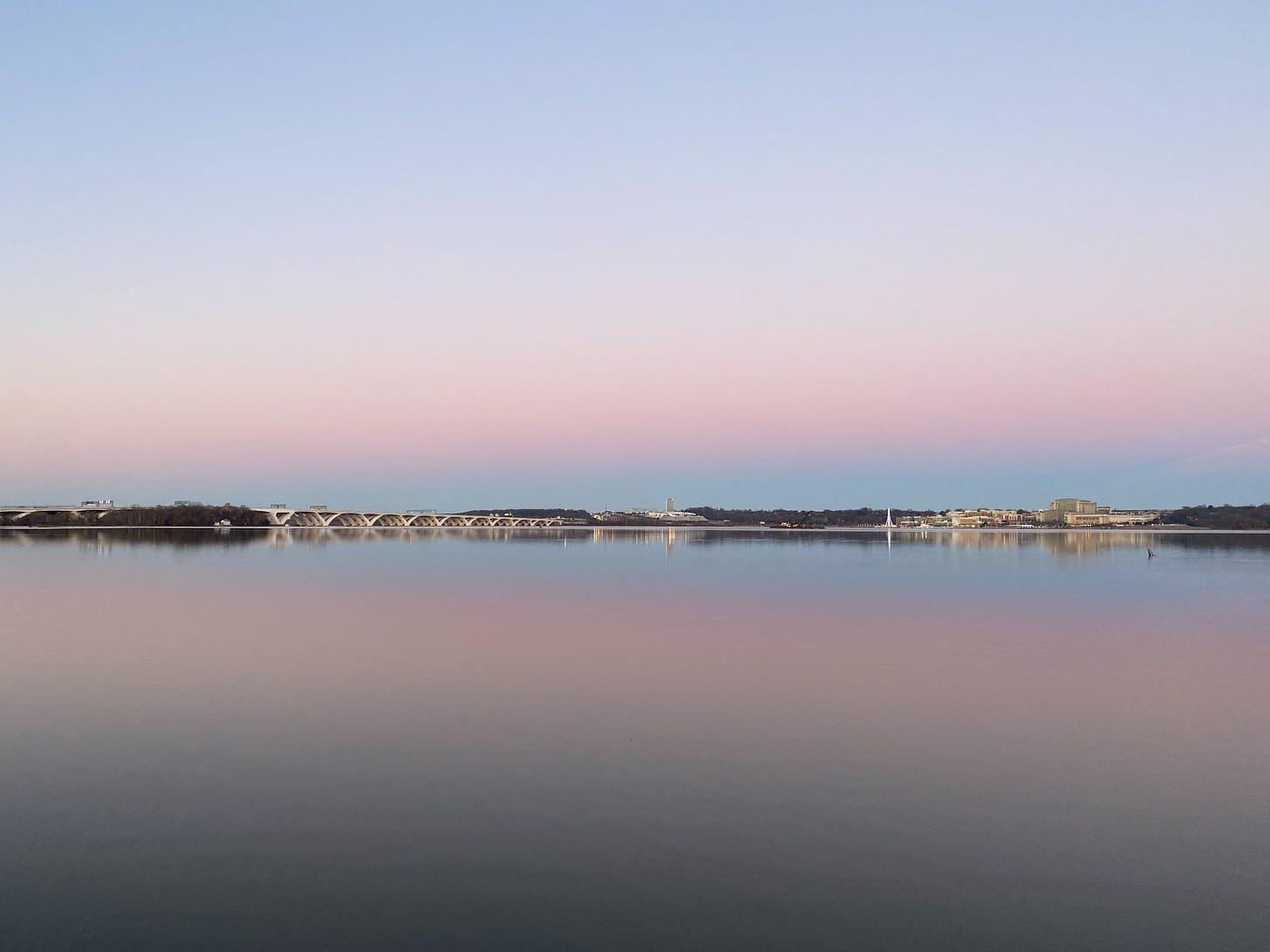 Photo of a calm, flat, wide river with a bridge in the distance and the sunset hues of blue, white, pink and purple reflected onto the water from the sky.