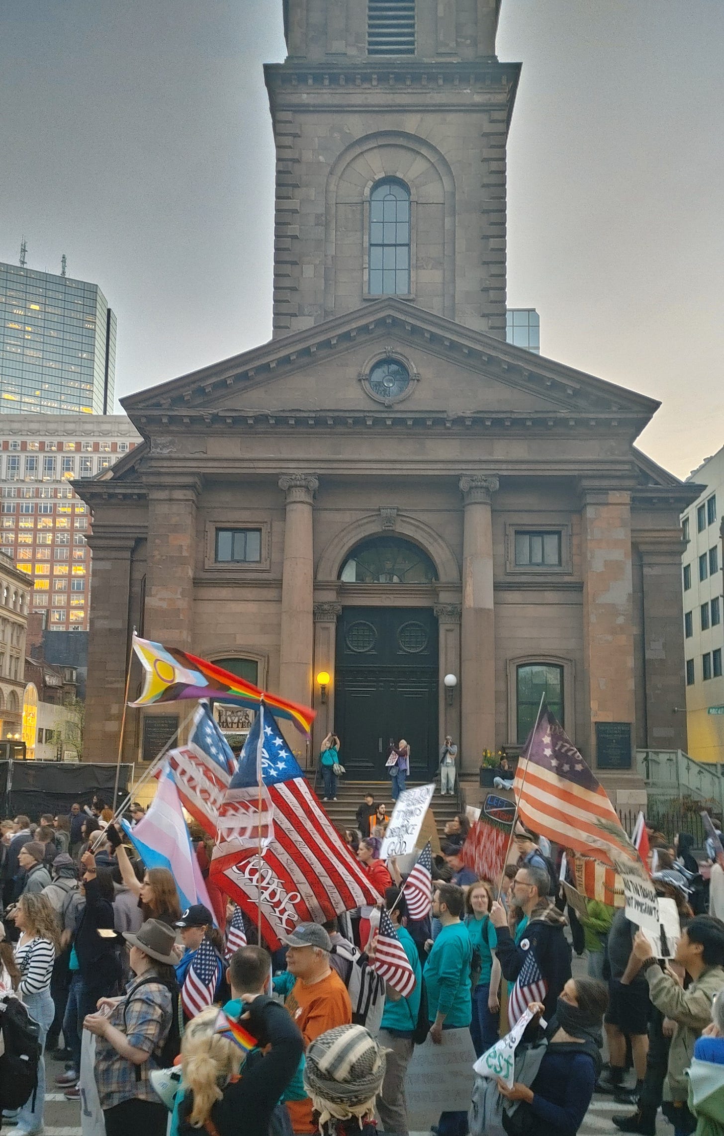 Protesters march in front of the Arlington Street Church in Boston. They are carrying signs and flags, some American, some American labeled "The Spirit of 76," some LGBTQIA pride, some Trans Pride. Protesters march in front of the Arlington Street Church in Boston. They are carrying signs and flags, some American, some American labeled "The Spirit of 76," some LGBTQIA pride, some Trans Pride.