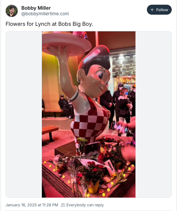 BlueSky post from Bobby Miller. Text: Flowers for Lynch at Bobs Big Boy. Image: The statue of Big Boy with flowers left at his feet. You can see the edges of the crowd of mourners gathered to remember David Lynch.