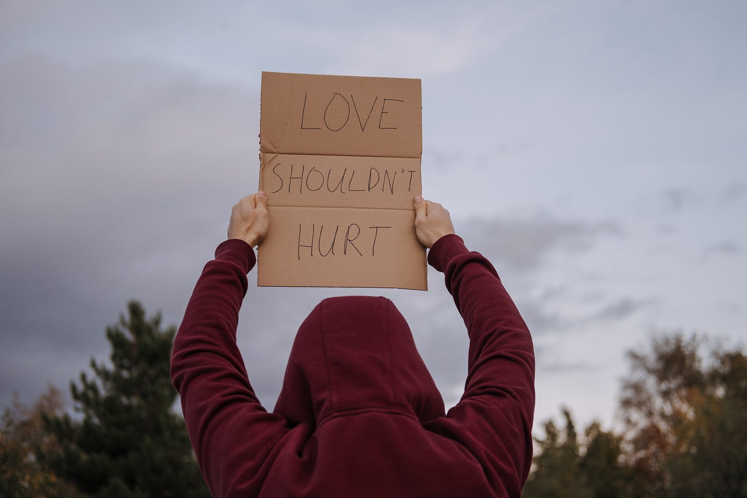 A person standing outside in a red hoodie, holding up a cardboard sign that says "love shouldn't hurt"