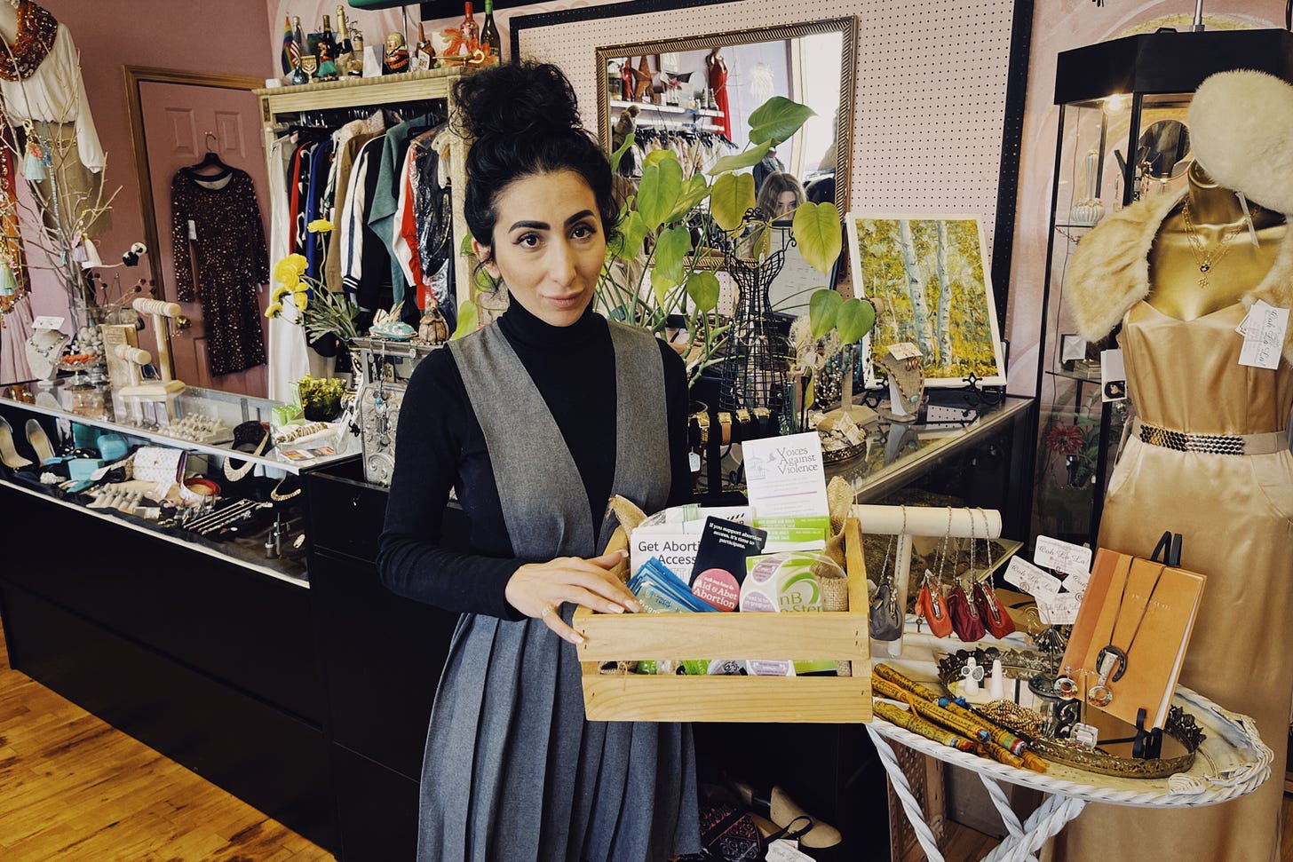 Liyah Babayan, an Armenian woman with black hair and wearing a black turtleneck with a gray dress over top, holds a reproductive health box containing emergency contraception, information about abortion, and more. Behind her is the checkout area of her boutique. It is a colorful space, with jewelry, clothing, and dress forms visible in the background.
