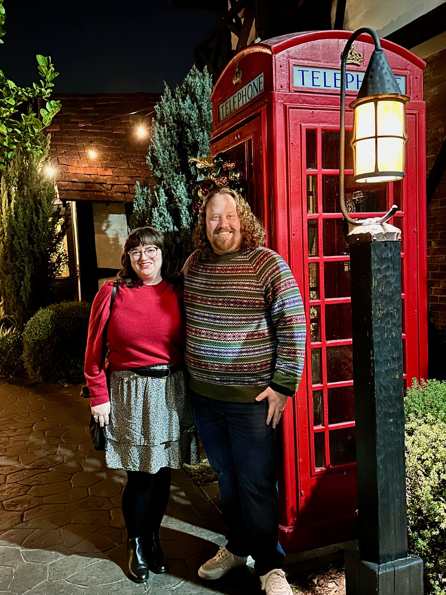 Photo at nighttime of Mike and me outdoors in front of a red telephone booth. I am wearing a red cashmere sweater, black and white skirt, black tights, and black boots. Mike is wearing a red white and green sweater, blue jeans, and white sneakers.