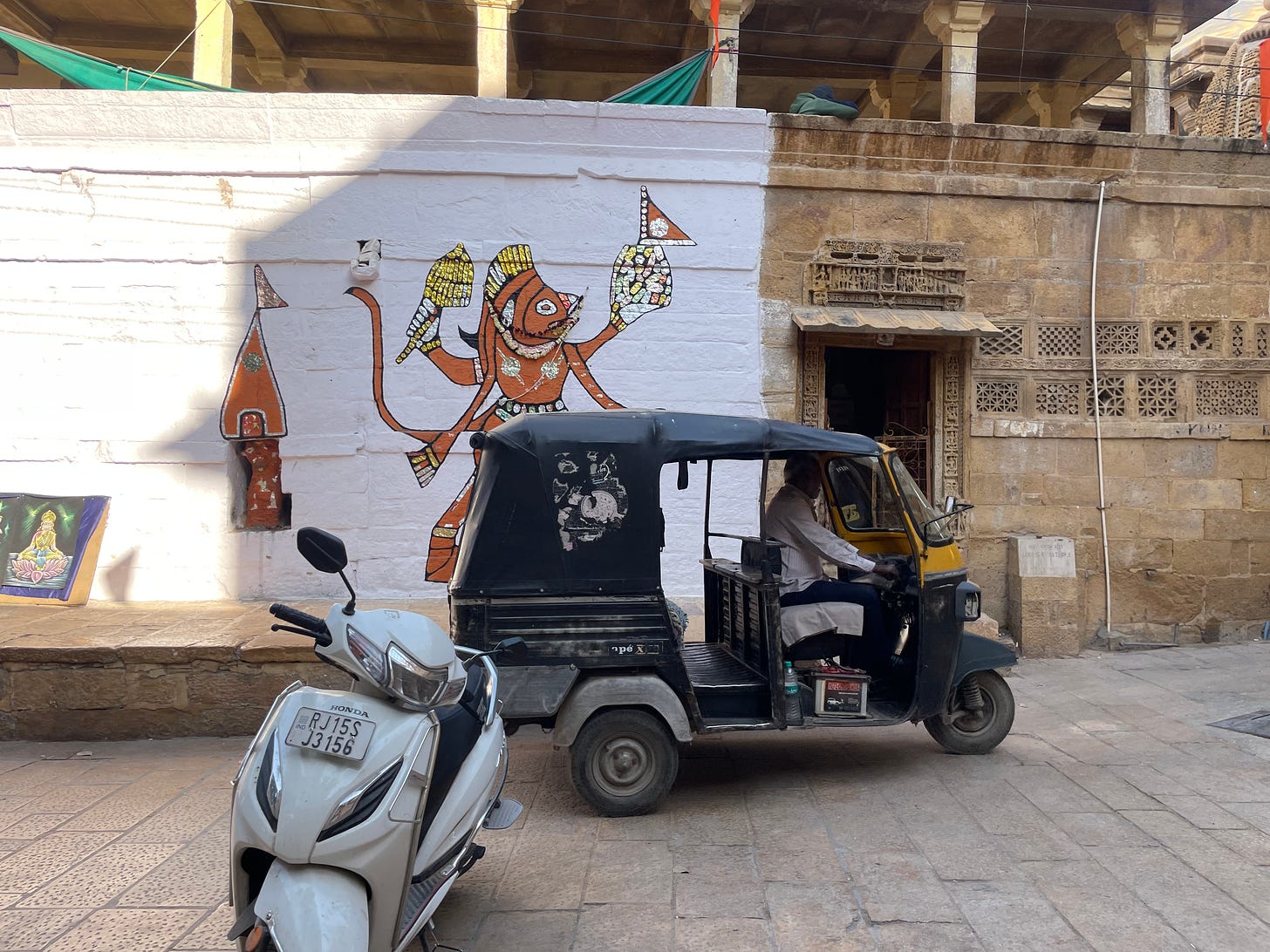 A driver sits in a rickshaw before a whitewashed wall painted with religious graffiti, and a sandstone wall with intricate carved windows.