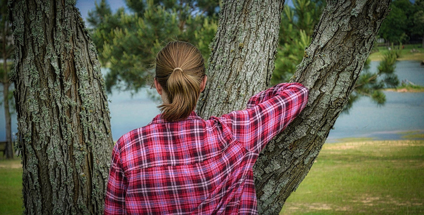 A young woman with a blonde pony tail wearing a pink plaid shirt looking out on a lake while leaning on the divided branches of a willow tree