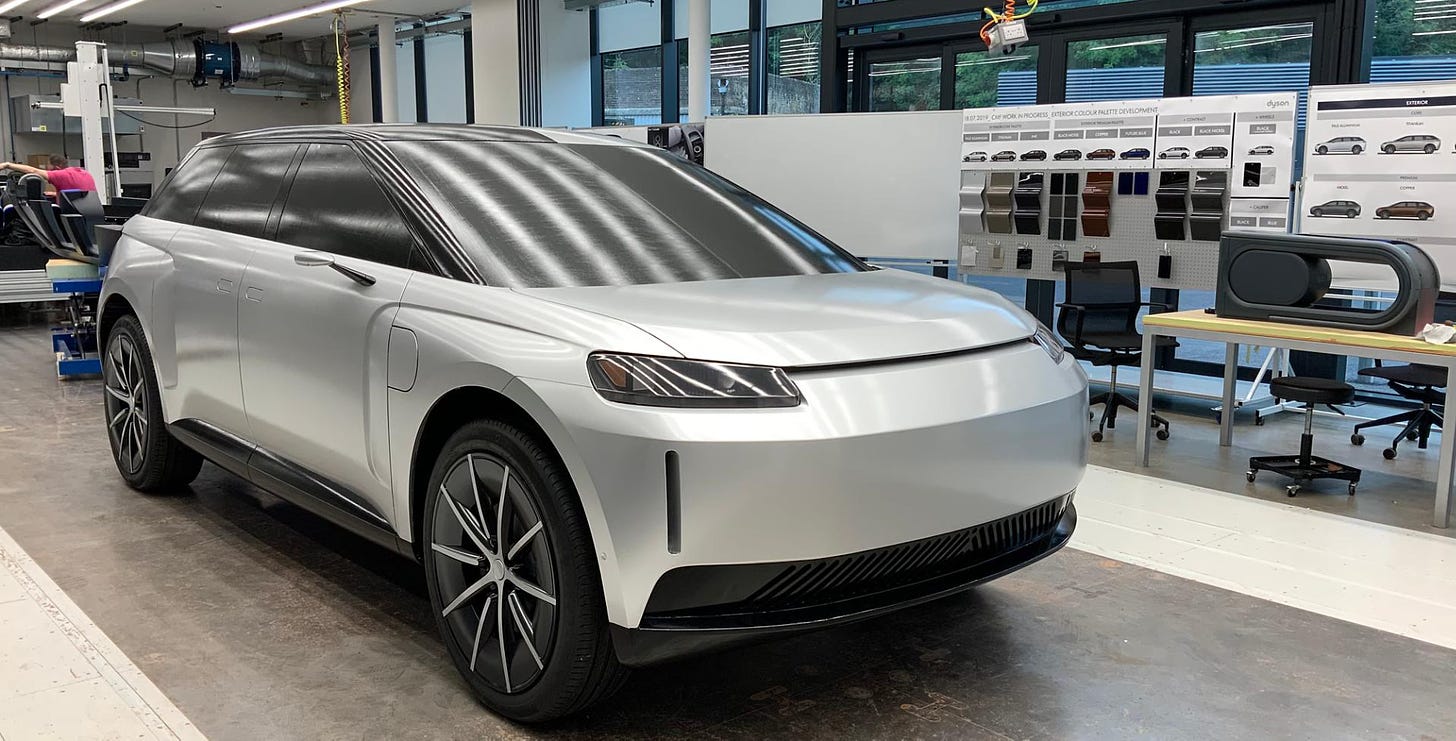 The image shows a white electric vehicle, likely a concept or prototype model. The vehicle has a sleek, futuristic design with sharp lines and an angular front grille. It appears to be an SUV or crossover-style vehicle with a high ground clearance and a large wheelbase. The interior of the vehicle is not visible, but the exterior design suggests advanced technology and an emphasis on aerodynamics and efficiency. The background indicates this is likely displayed in a showroom or design studio setting, with various product information boards visible.