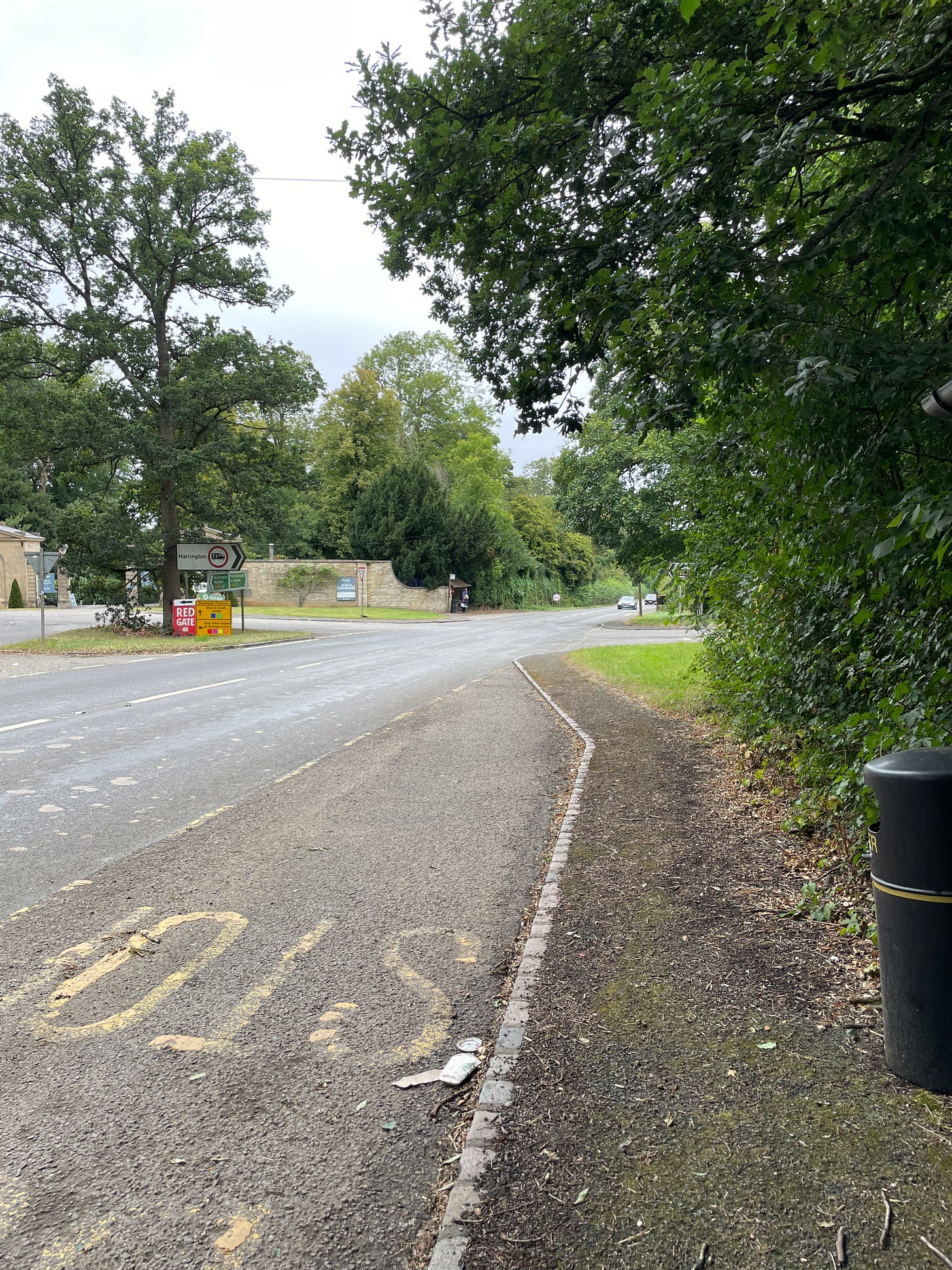 An a-road with a bus sign makred on the ground. Opposite is a stately home and another bus stop with a person with a big backpack waiting at it.