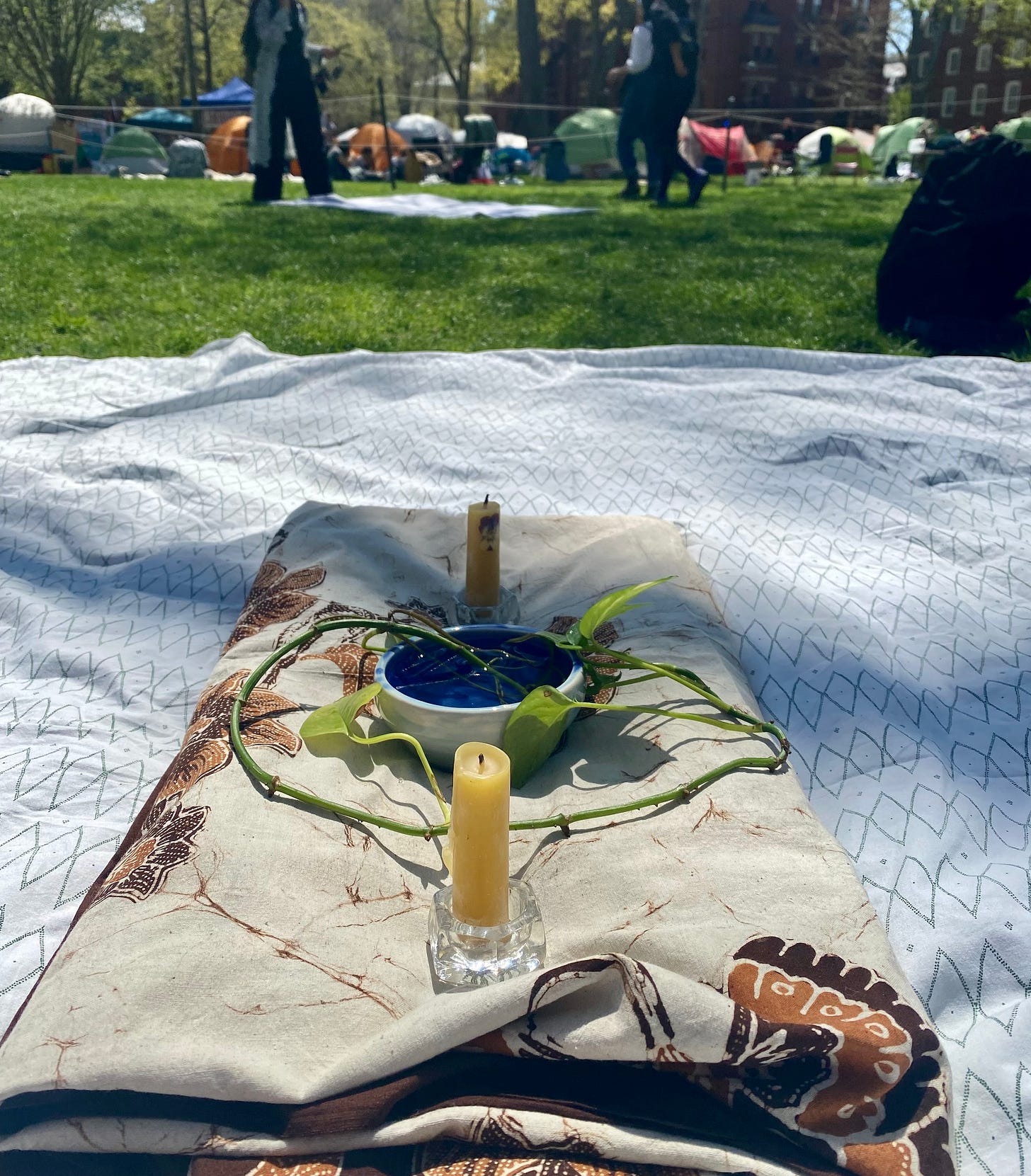 Folded blanket atop a sheet, with a plant and candles. Background of a campus green with tents in the background.