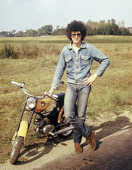 A guy by the side of the road near a field in Hungary. He's wearing a jean jacket and standing next to a a motor bike. He has big curly hair.