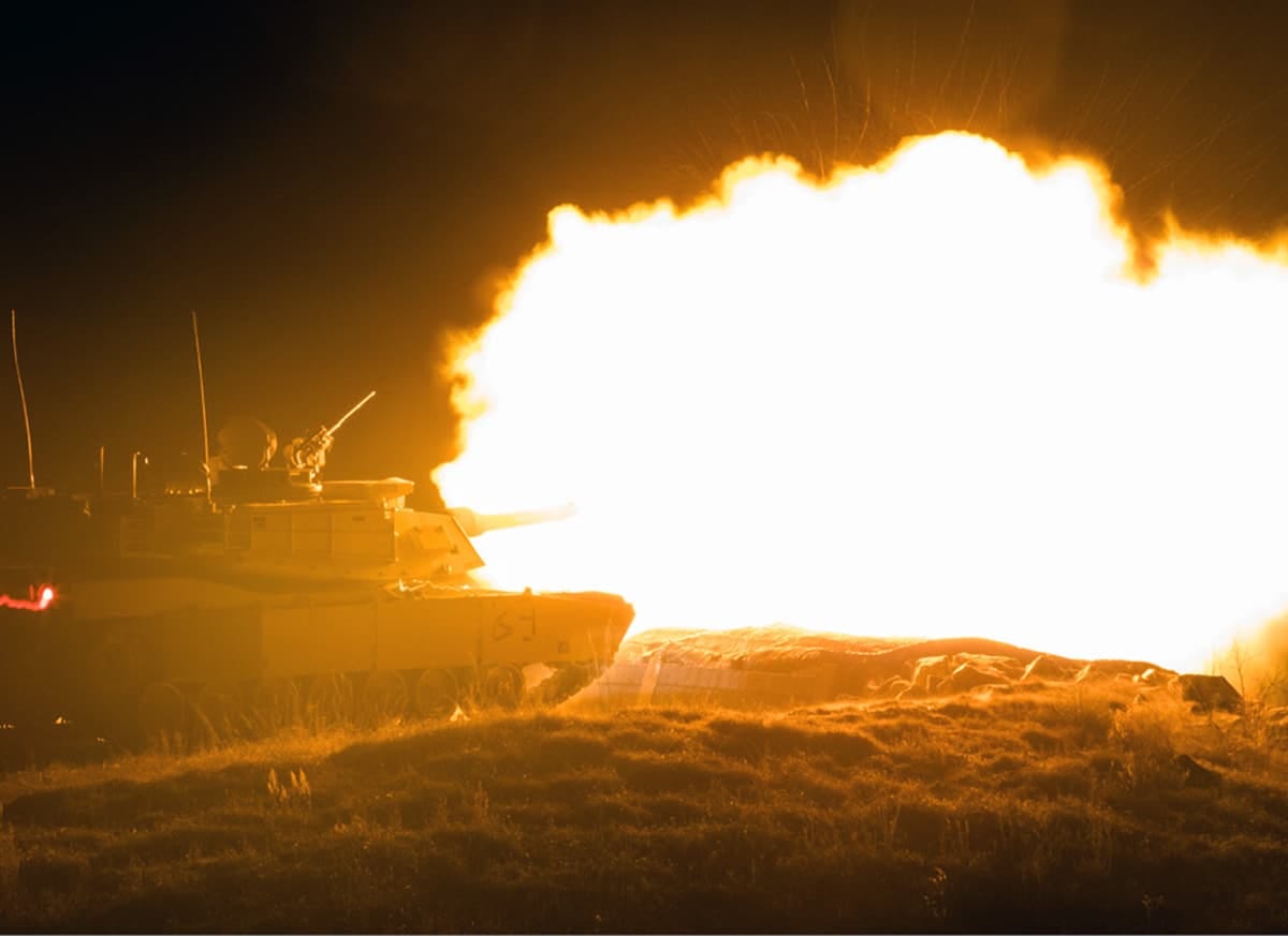 An M1 Abrams tank fires its main gun at night, creating a large fireball and plume of smoke.