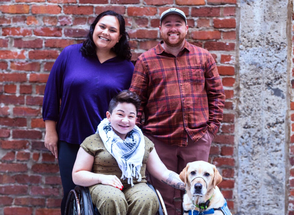 A multi-racial disabled family and a service dog smile for their portrait against a red brick wall outside.