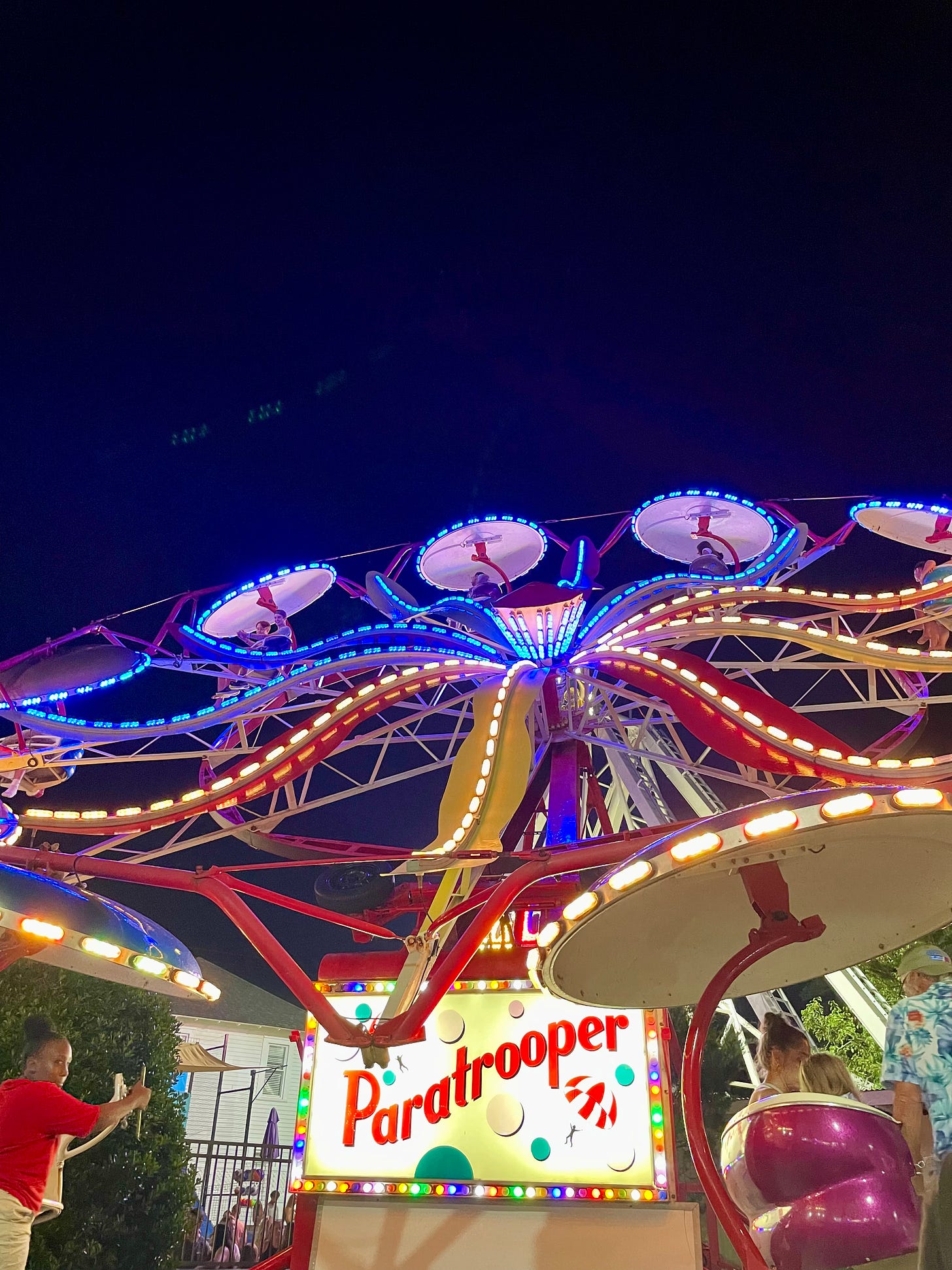 Brightly lit carnival ride against a night sky. The ride is like a ferris wheel, with people dangling from blue-illuminated disks, circling a wheel. The words "Paratrooper" are at the bottom of the ride.