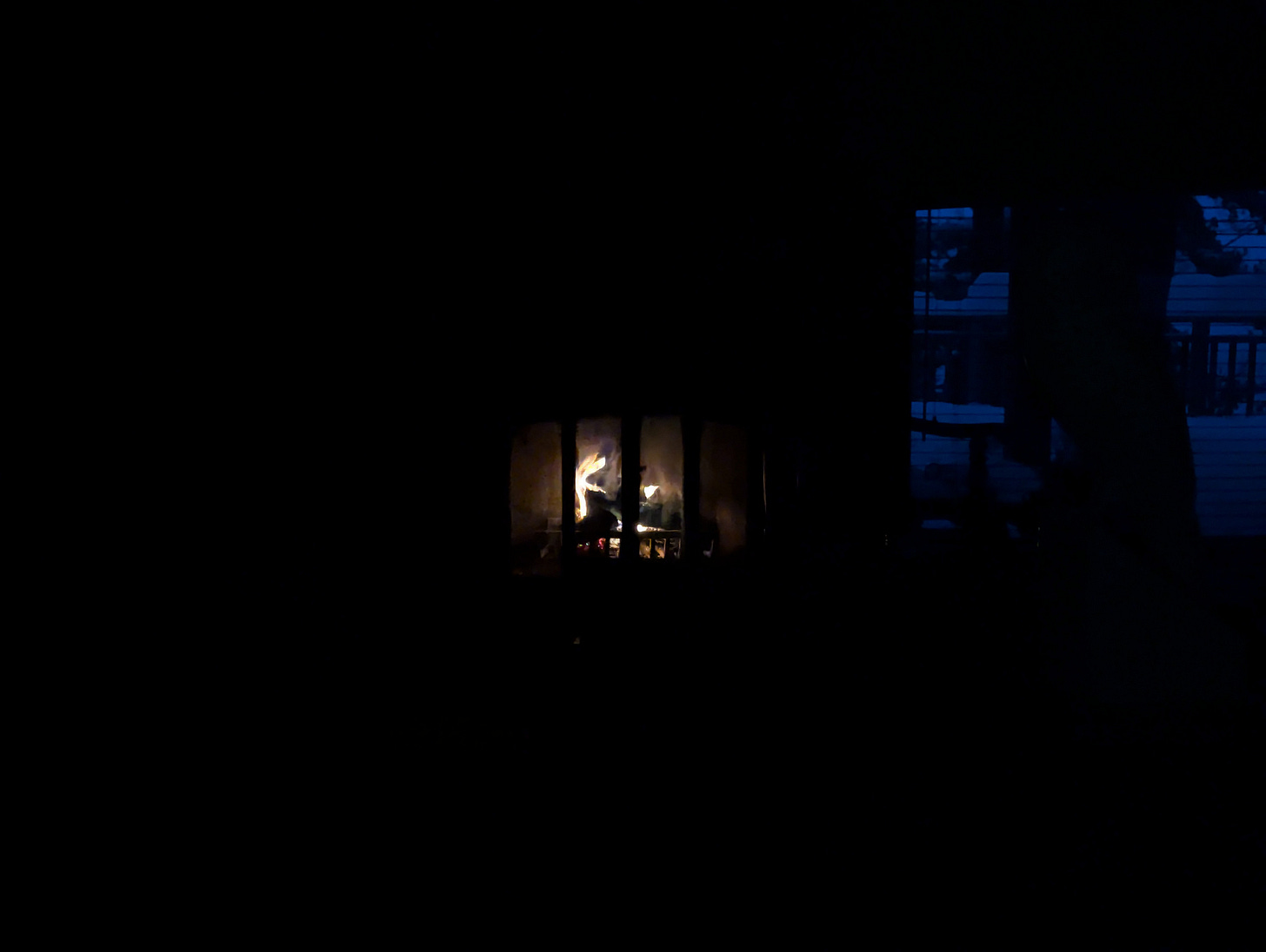 An open fireplace at Aleksa's cottage burns, surrounded by pitch black, with the dusk appearing in the window through blinds on the right.