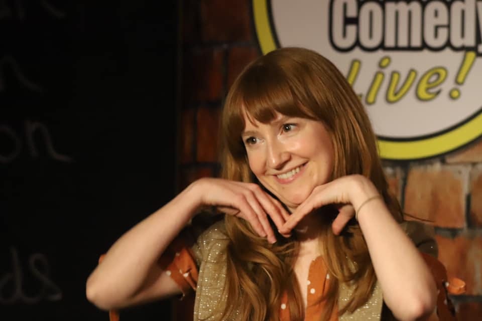 Woman with long ginger hair smiles inanely with hands cupping her face. She is on a stage with comedy logo out of focus in background