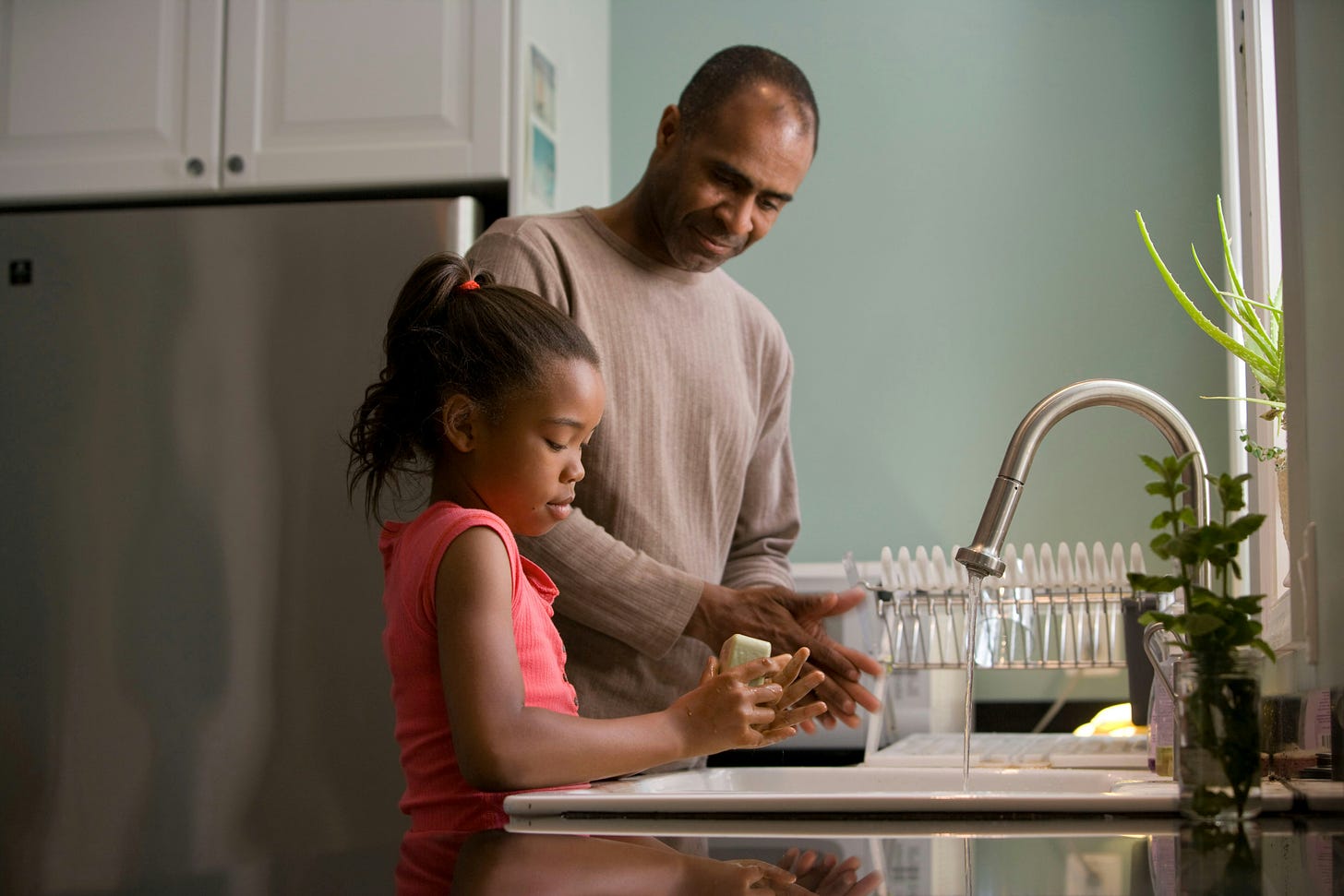 A Black father and daughter washing hands at a kitchen sink.