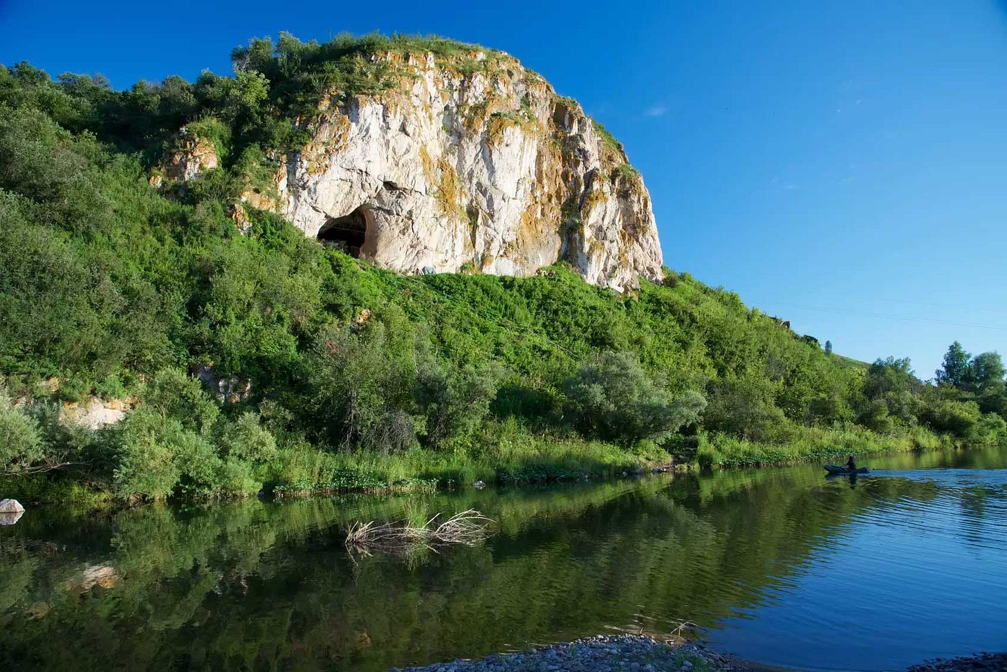 Chagyrskaya Cave in a sheer limestone cliff overlooking a river with blue sky Chagyrskaya Cave in a sheer limestone cliff overlooking a river with blue sky