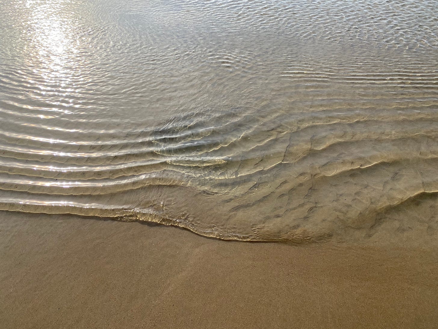 Sunlit shallow water on a sandy beach, ripples and a deeper hole under the water.