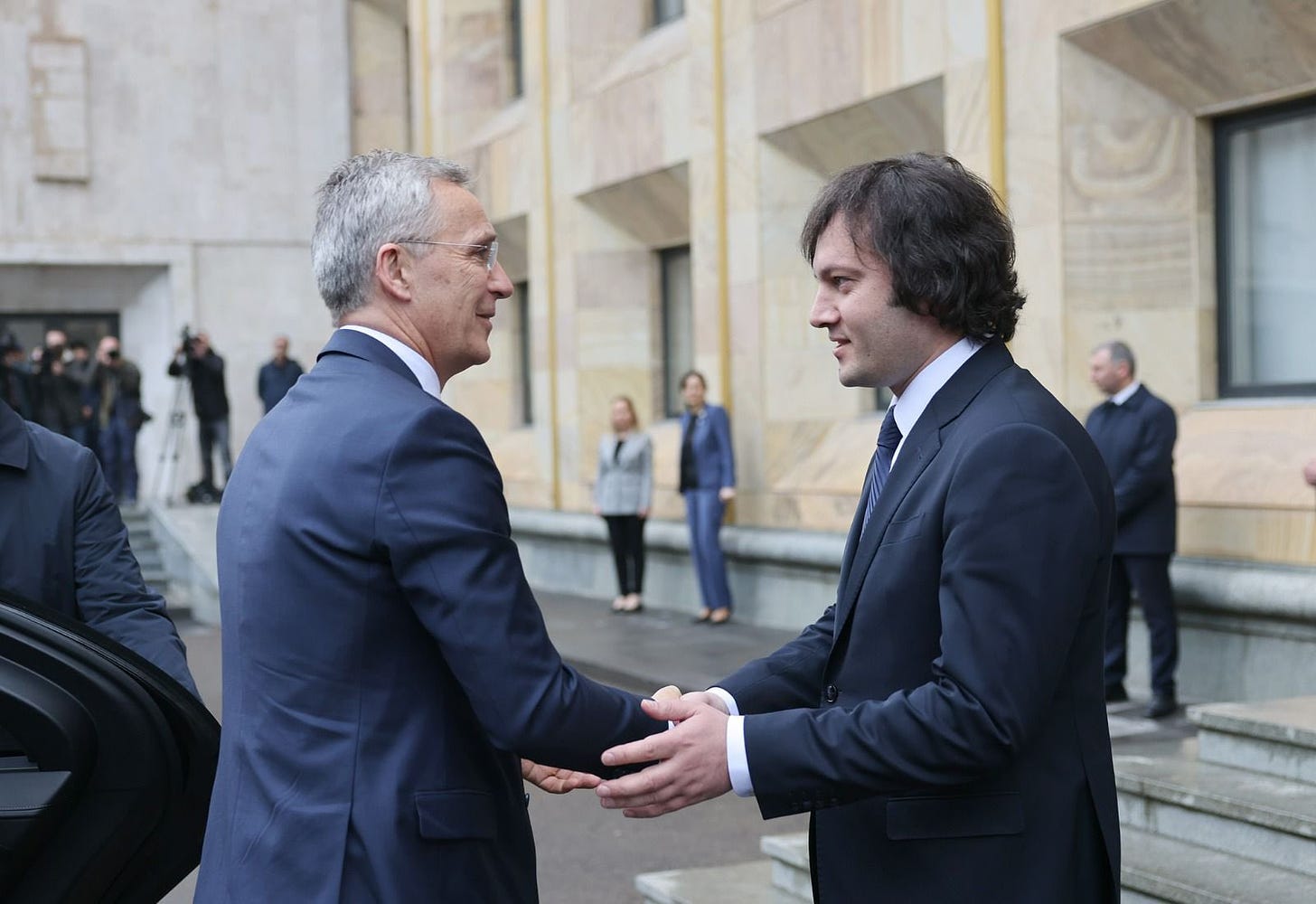 A person in suit shaking hands with another person in the background

Description automatically generated