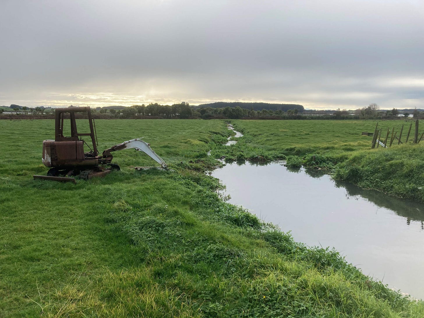 A polluted stream running through a green field, with a digger beside it and misty light in the distance