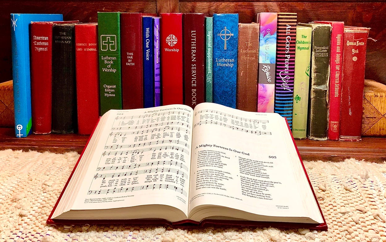 A variety of service books and hymnals sit in a row on a shelf. Another hymnal is open to A Mighty Fortress is Our God in front of this shelf.