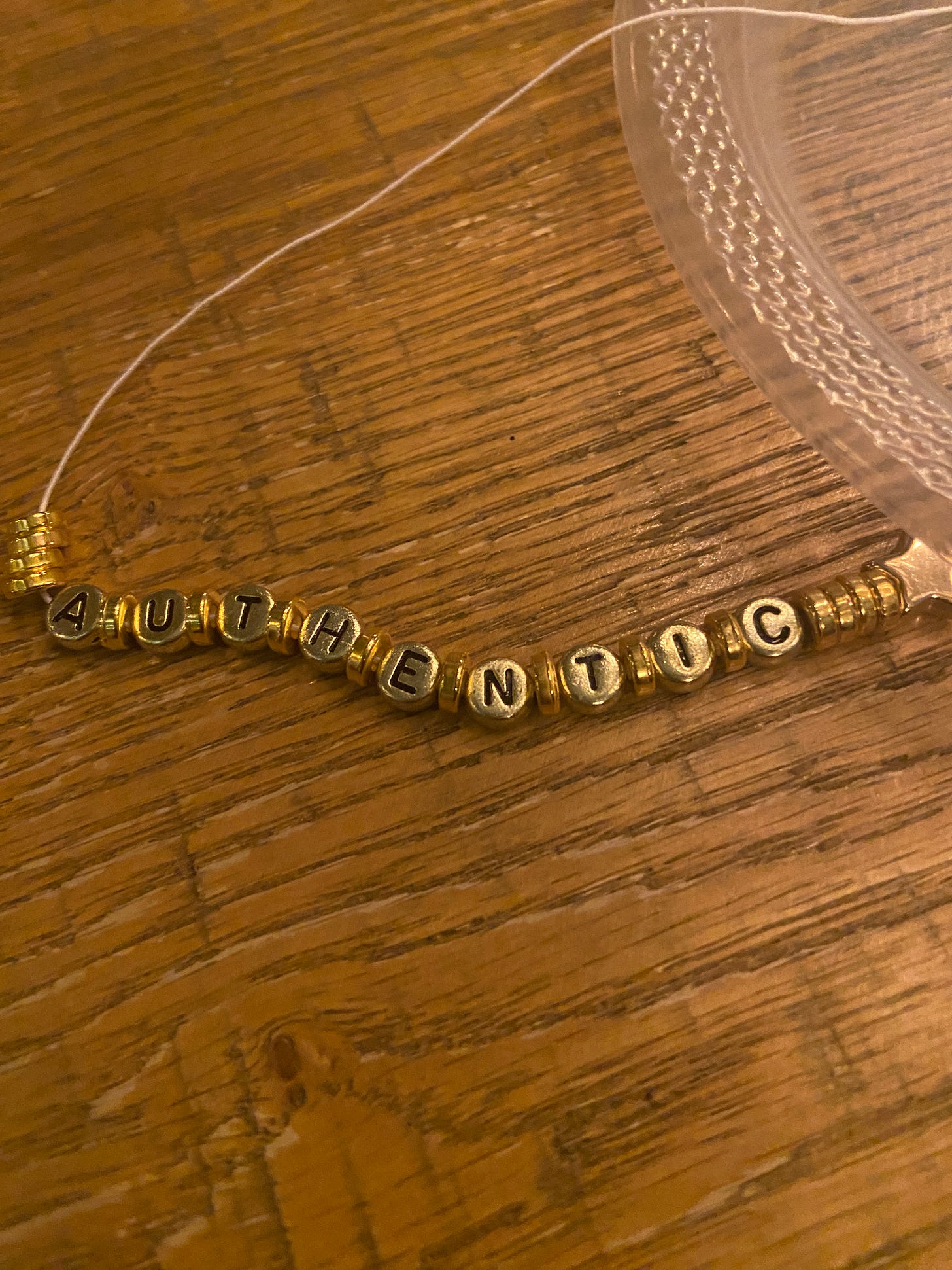 a beaded braclet with a few round gold beads, and letters spelling out AUTHENTIC with beads betweent the letters. There is also a star shaped bead on the far right of the bracelet. Some of the white string is showing on the left side of the letters. The bracelet is laying flat, straight, and horizontal (before being tied togerther) on a brown wooden table. You can see the corner of a plastic plate in the upper right hand side of the photo.