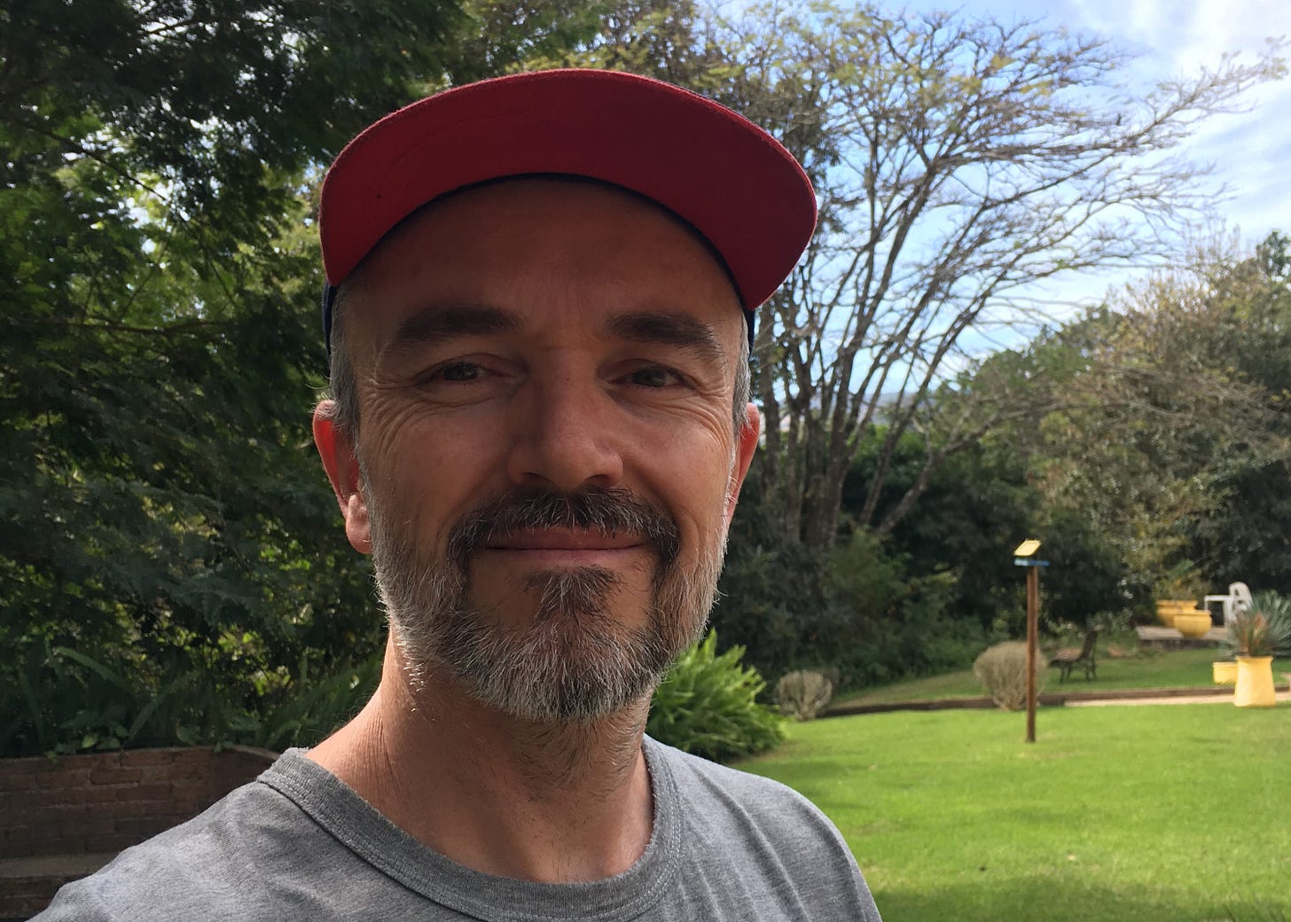 A middle-aged man in a garden, with a grey T-shirt and a blue and red baseball cap, smiling at the camera. He has a light salt-and-pepper beard and he's smiling.