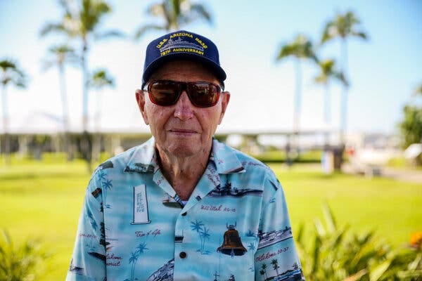 A close-up color photo of Mr. Potts wearing a blue U.S.S. Arizona cap, sunglasses and a blue open-collared shirt printed with images of palm trees, a bell and Navy ships. Palm trees standing in sunlight are behind him.