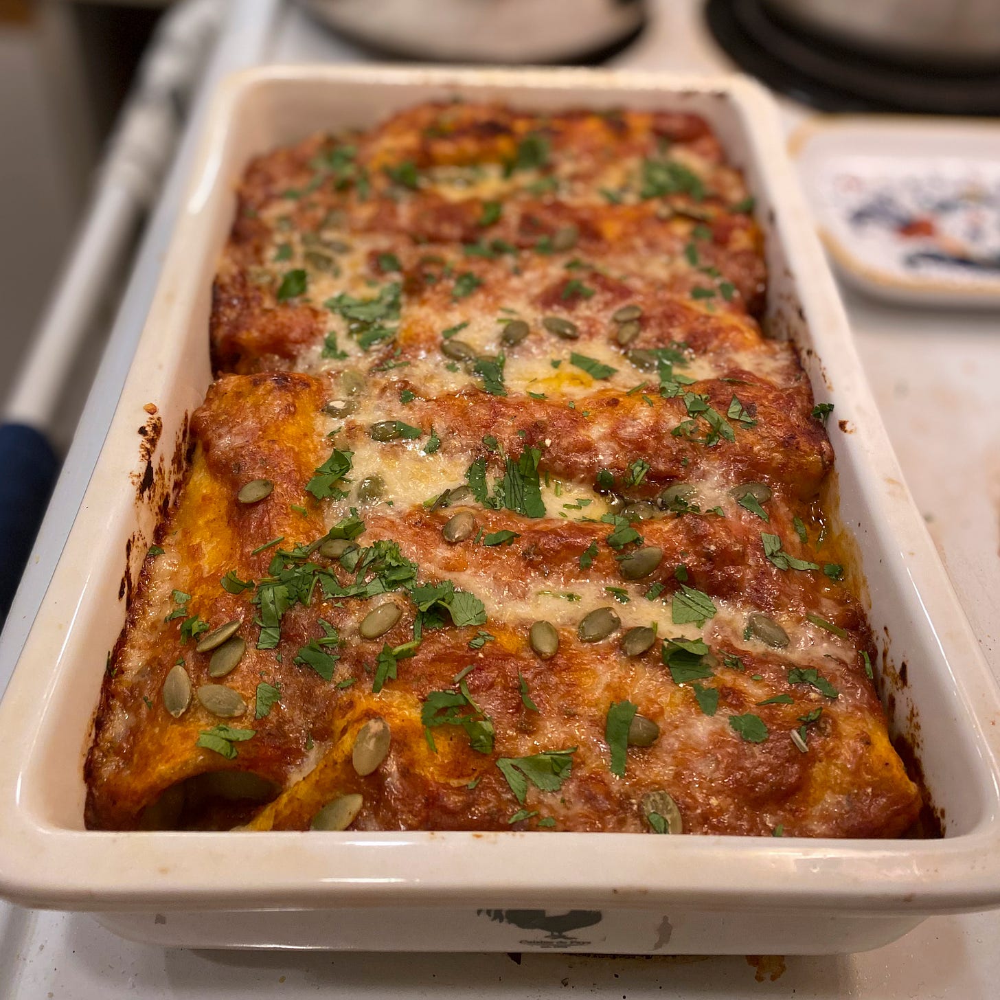 A white ceramic baking dish of the enchiladas. Sauce has caramelized around the edges and they are covered in melted white cheddar, a sprinkling of cilantro, and pumpkin seeds.
