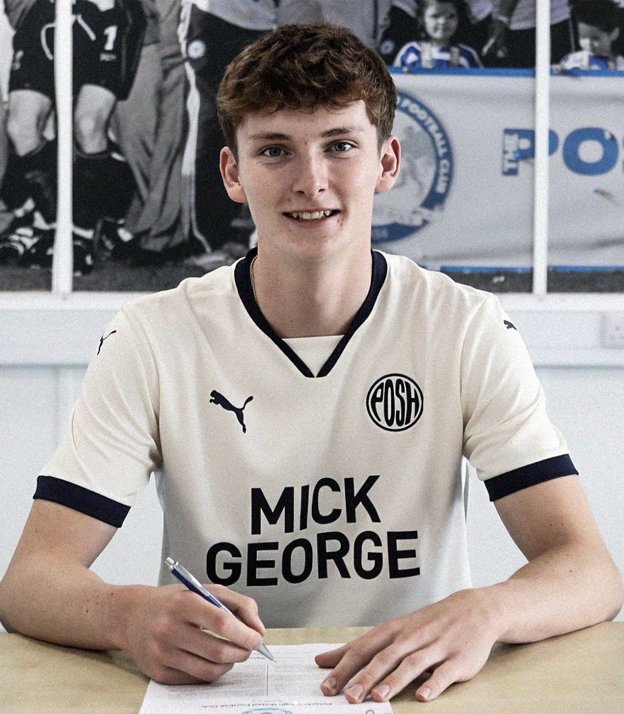 A photo of George Nevett wearing a retro-inspired white Peterborough United shirt, sat at a table with a pen in his right hand and a contract in front of him