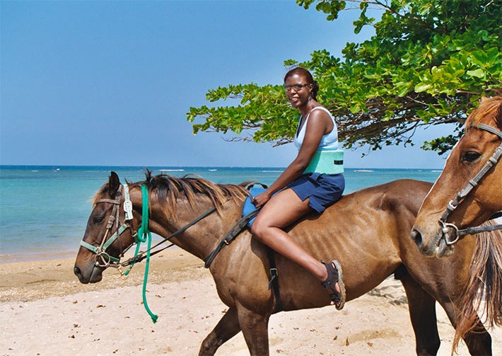 At the beach in Jamaica with blue sky and turquoise sea in the background. Sandy shoreline, Tanya Owens on a horse with a green bush behind her and another horse's face in the foreground