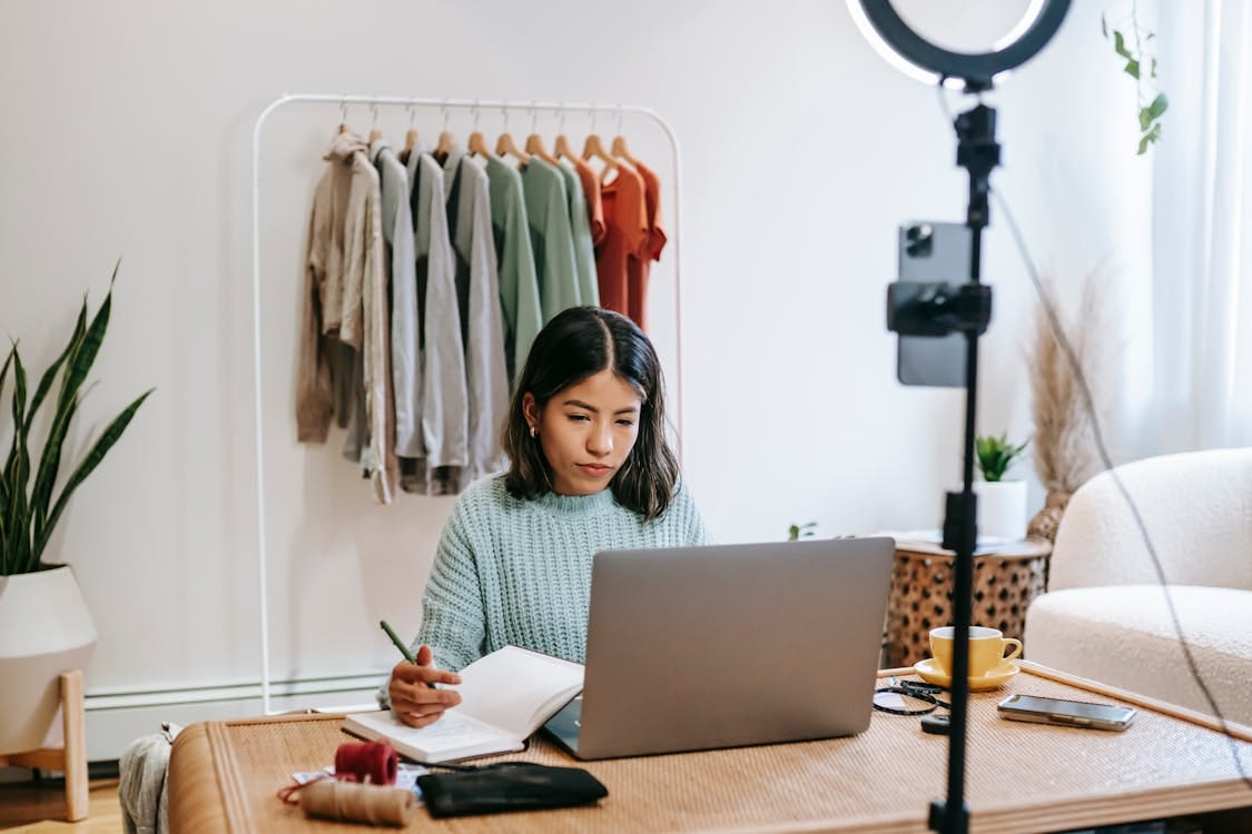 Free Focused young woman working on laptop in home office, surrounded by clothing rack and camera setup. Stock Photo