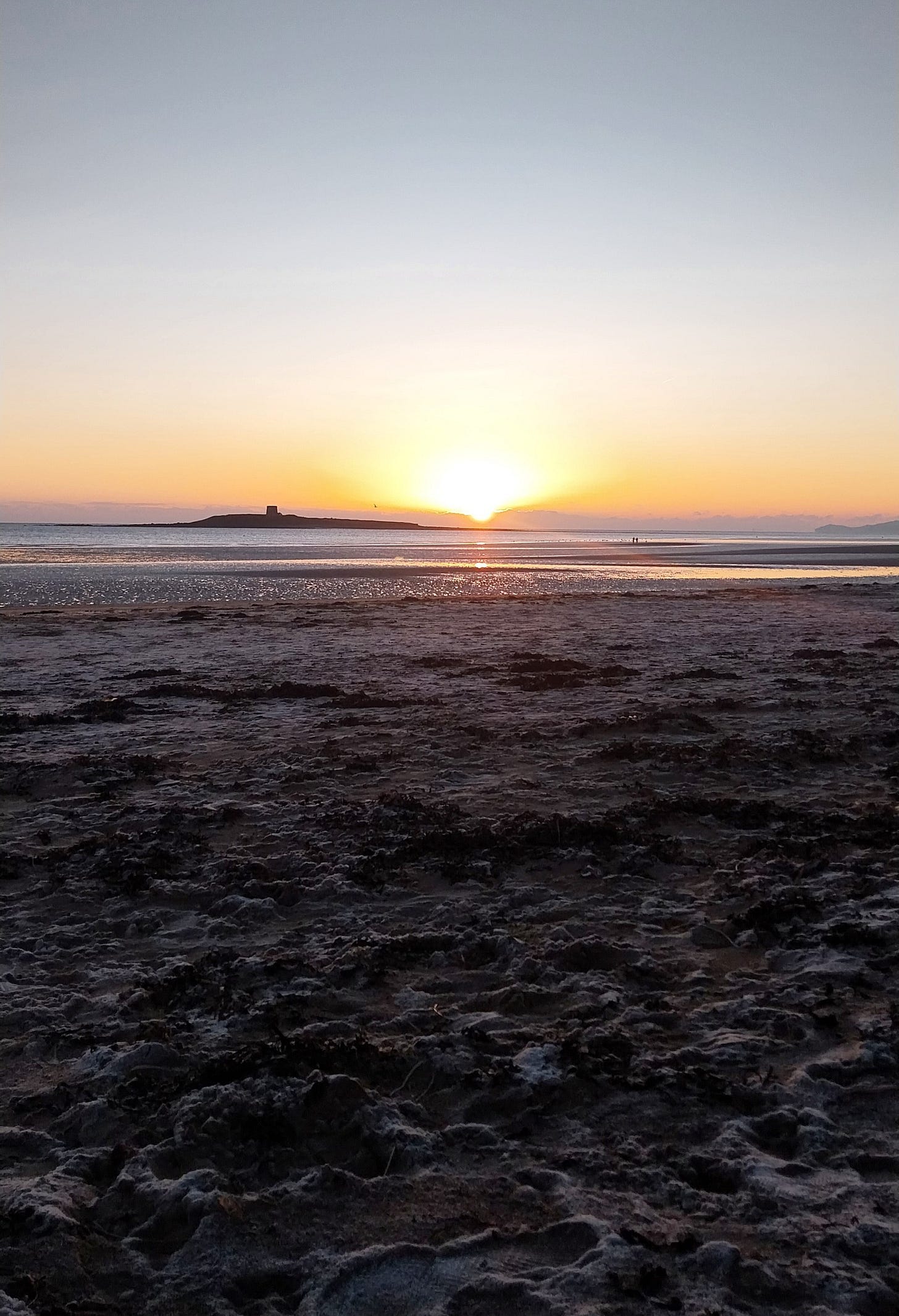 Sunrise over Skerries beach at low tide, with golden light breaking over a small island, casting rays across the frosty sand.