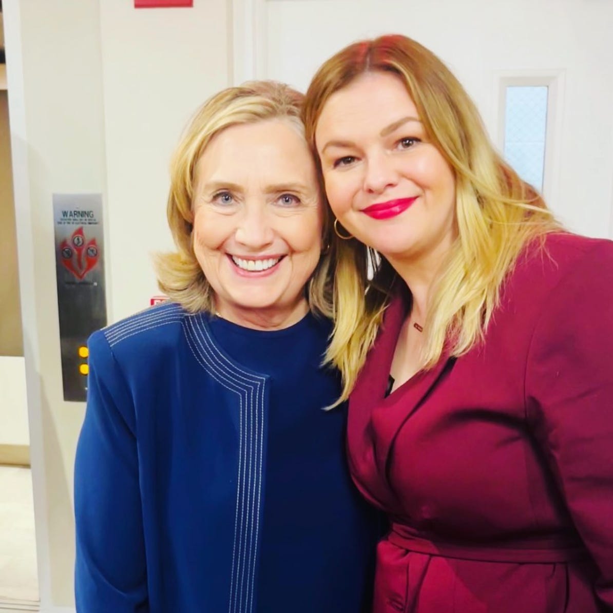 Hillary Clinton and Amber Tamblyn stand with their arms around each other and their heads touching. They both smile into the camera. Hillary wears a blue top and jacket. Amber wears a fuchsia jacket.