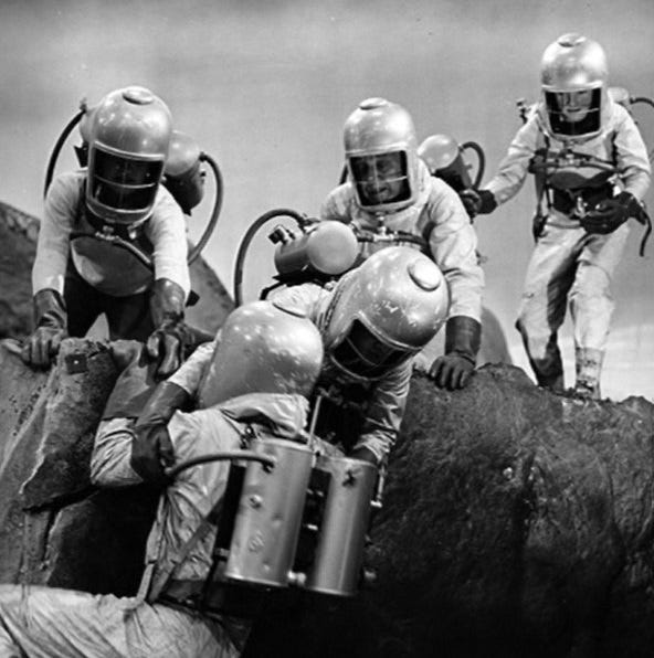 Four characters in spacesuits upon a rocky ledge. One of them is aiding a fourth spacesuited character who is struggling to cling on to the ledge.