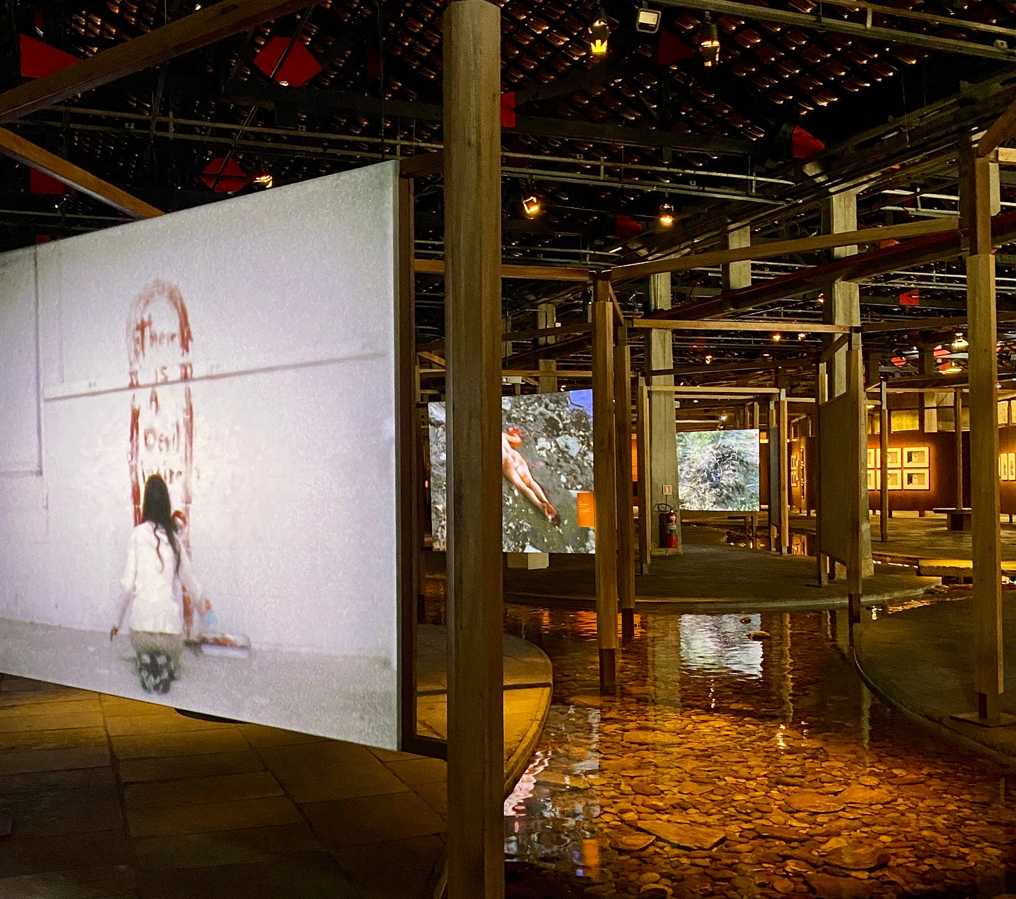 Imagem: Espaço expositivo do Sesc Pompeia com diversos telões com vídeos sendo projetados e um corrego de agua entre eles. Em volta estruturas de madeiras para separar cada telao.