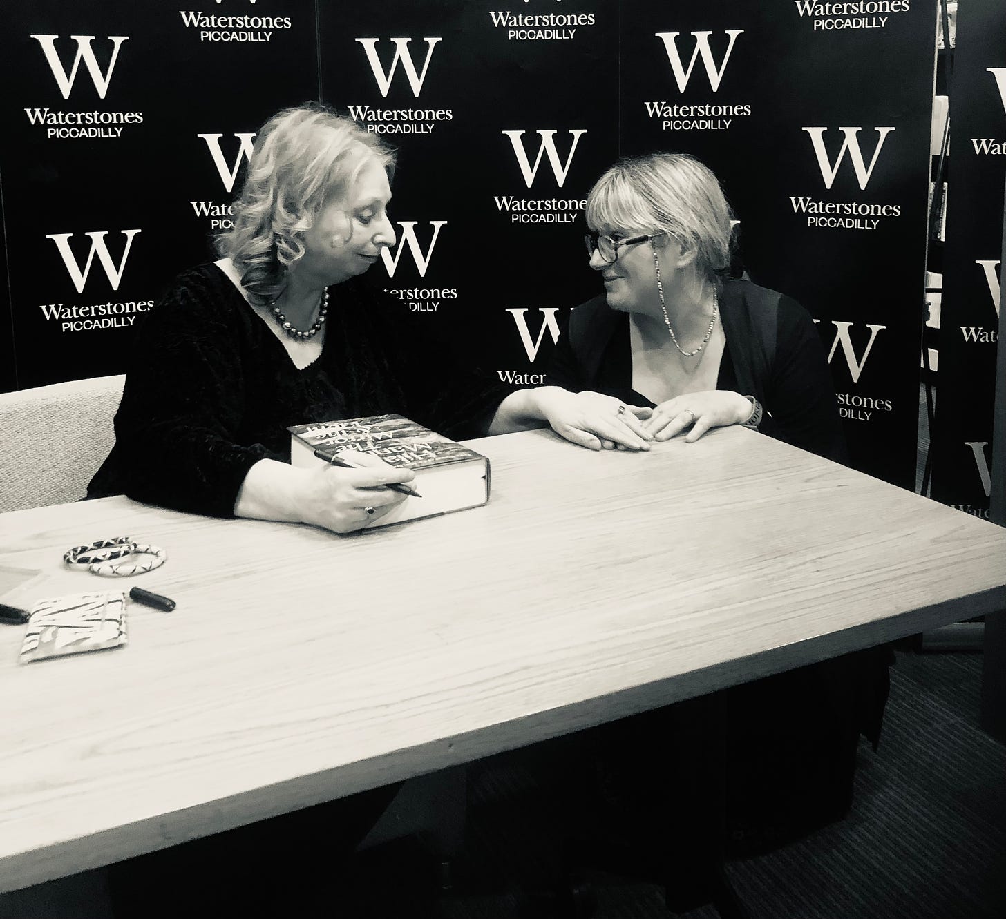 Two women at a table. One woman has her hand on the other’s; and is holding a book and a pen.
