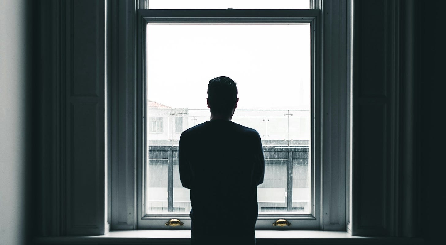 The silhouette of a man standing in front of a bright window. There is a stark constrast between the darkness of the room he is in and the light outside.
