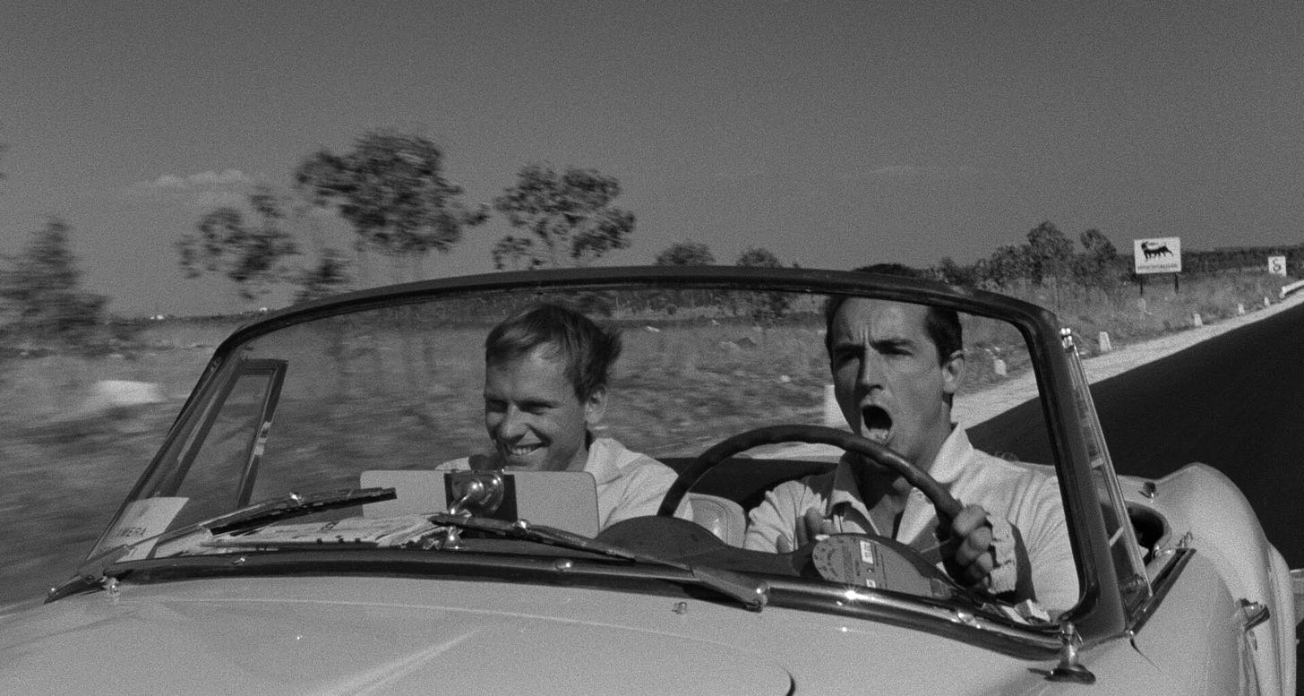 A black and white image from the movie Il Sorpasso. We see two men in a car driving in the Italian countryside. The driver is singing.